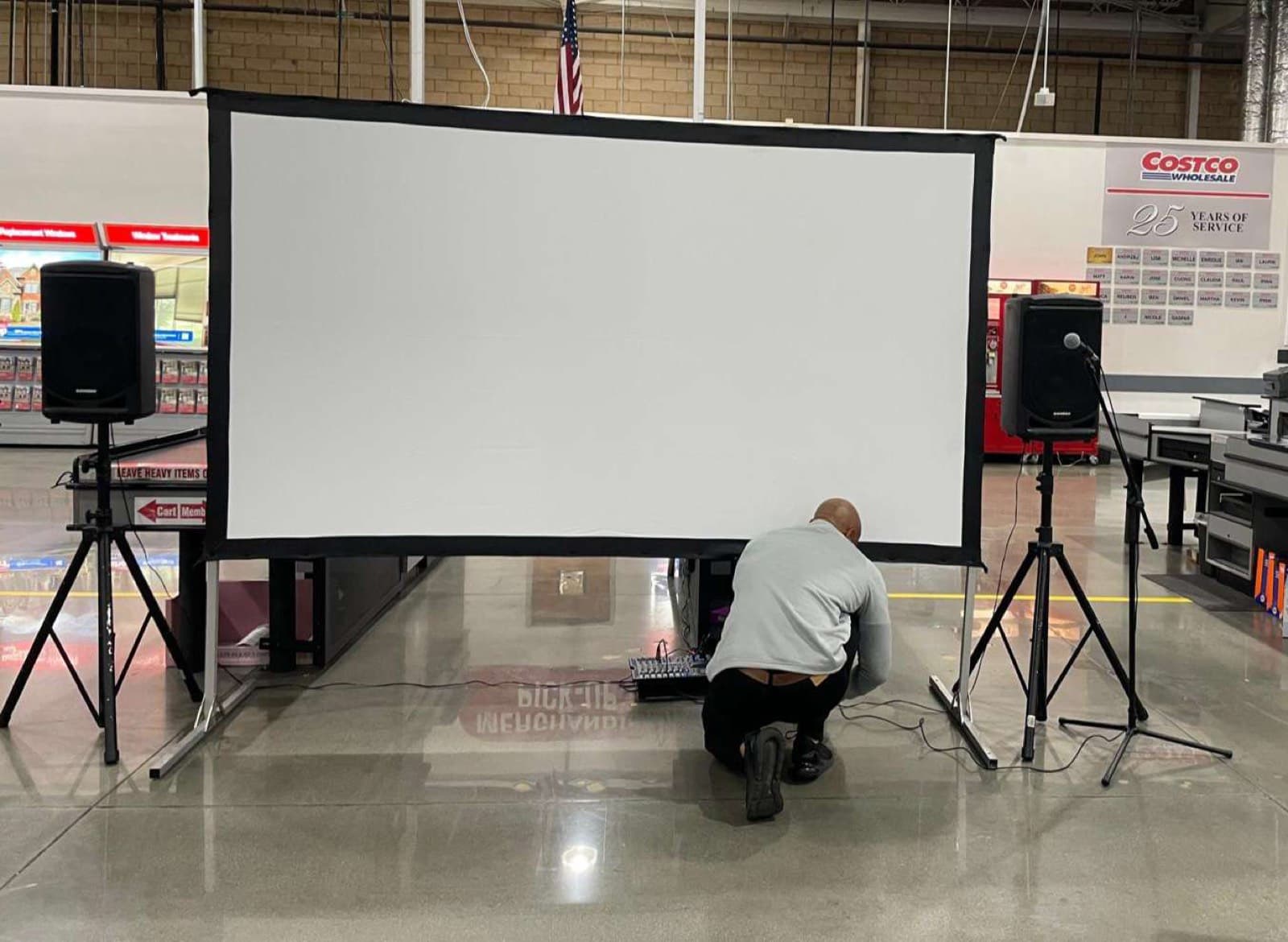 Corporate event AV setup at a Costco warehouse — large fast-fold projection screen, PA speakers, and audio mixer